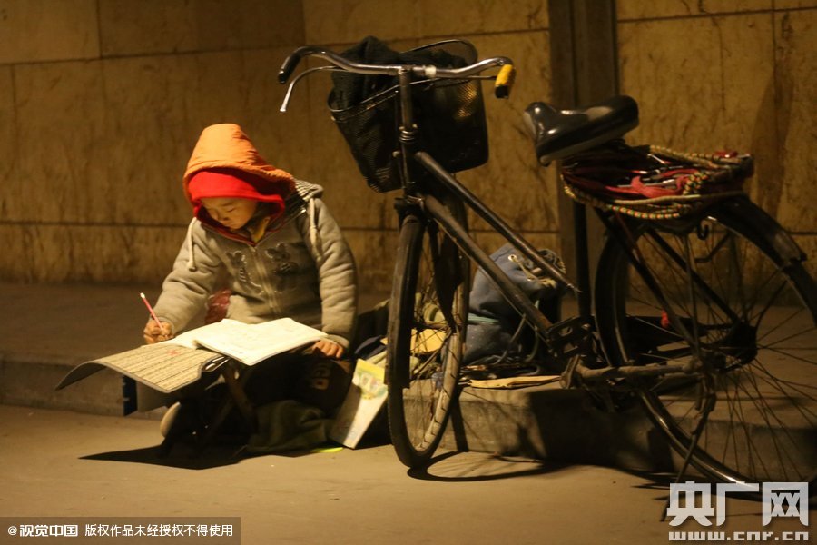 鄭州中原路京廣鐵路大橋下涵洞的馬路牙子邊，一位戴著紅色辣椒帽的小男孩趴在一個小馬扎上，上面鋪著一張車墊，在微弱的燈光下寫作業(yè)